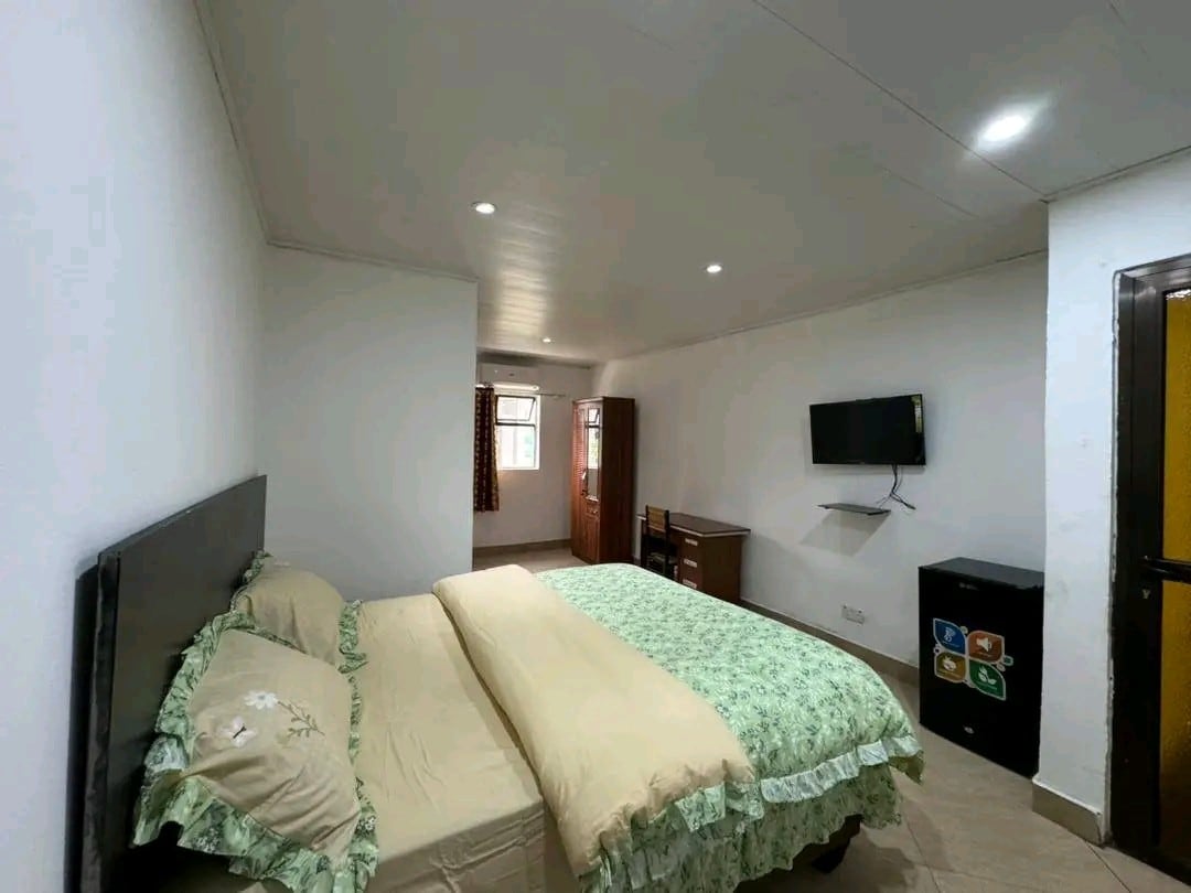 A well-lit room features a spacious bed dressed in soft green linens and an elegant floral trim. A wooden desk is positioned nearby, with a wall-mounted television providing entertainment. Natural light filters in through a window, complemented by light-colored walls that enhance the bright atmosphere.