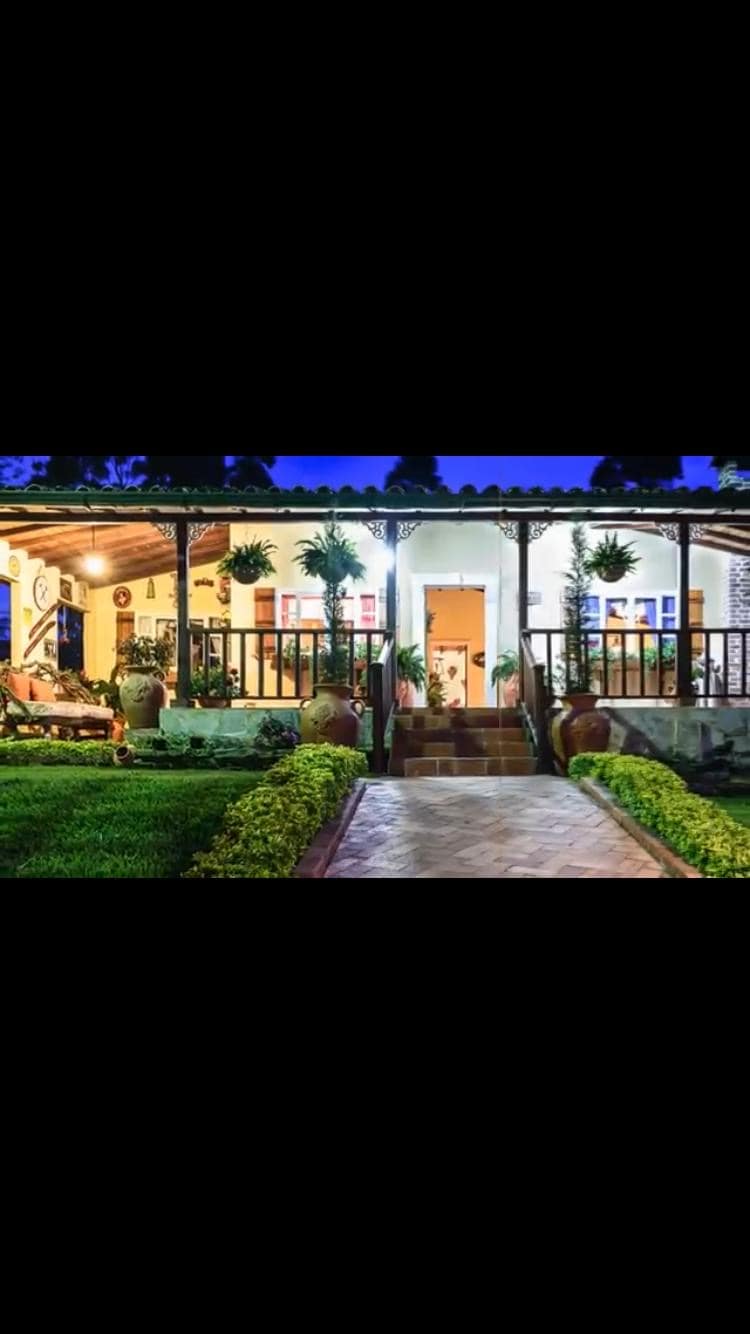 The exterior of the cottage features an inviting porch adorned with hanging plants. A pathway leads to a wooden door framed by greenery, while well-maintained lawns and vibrant flowering shrubs create a serene entrance to this picturesque hideaway.