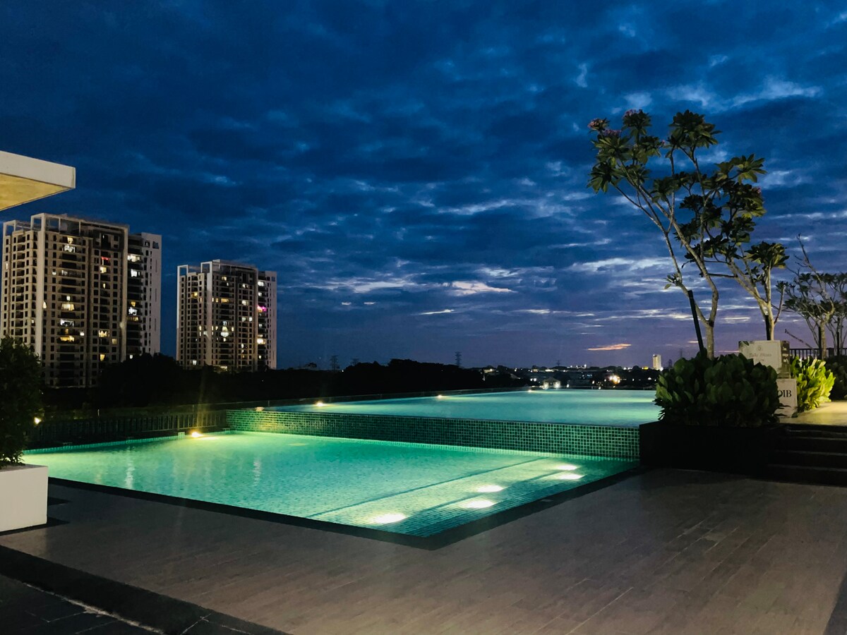 An inviting pool area is illuminated under a twilight sky, featuring two expansive swimming pools. Lush greenery surrounds the space, and modern residential buildings are visible in the background. Soft lights reflect on the water's surface, creating a calming ambiance.