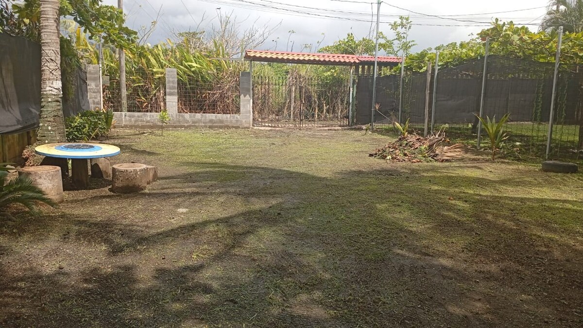 A spacious outdoor area is surrounded by greenery, featuring a small circular table and seating made from natural materials. The ground is covered with grass, and a shaded section is observed, while a fence and a roofed structure are visible in the background.