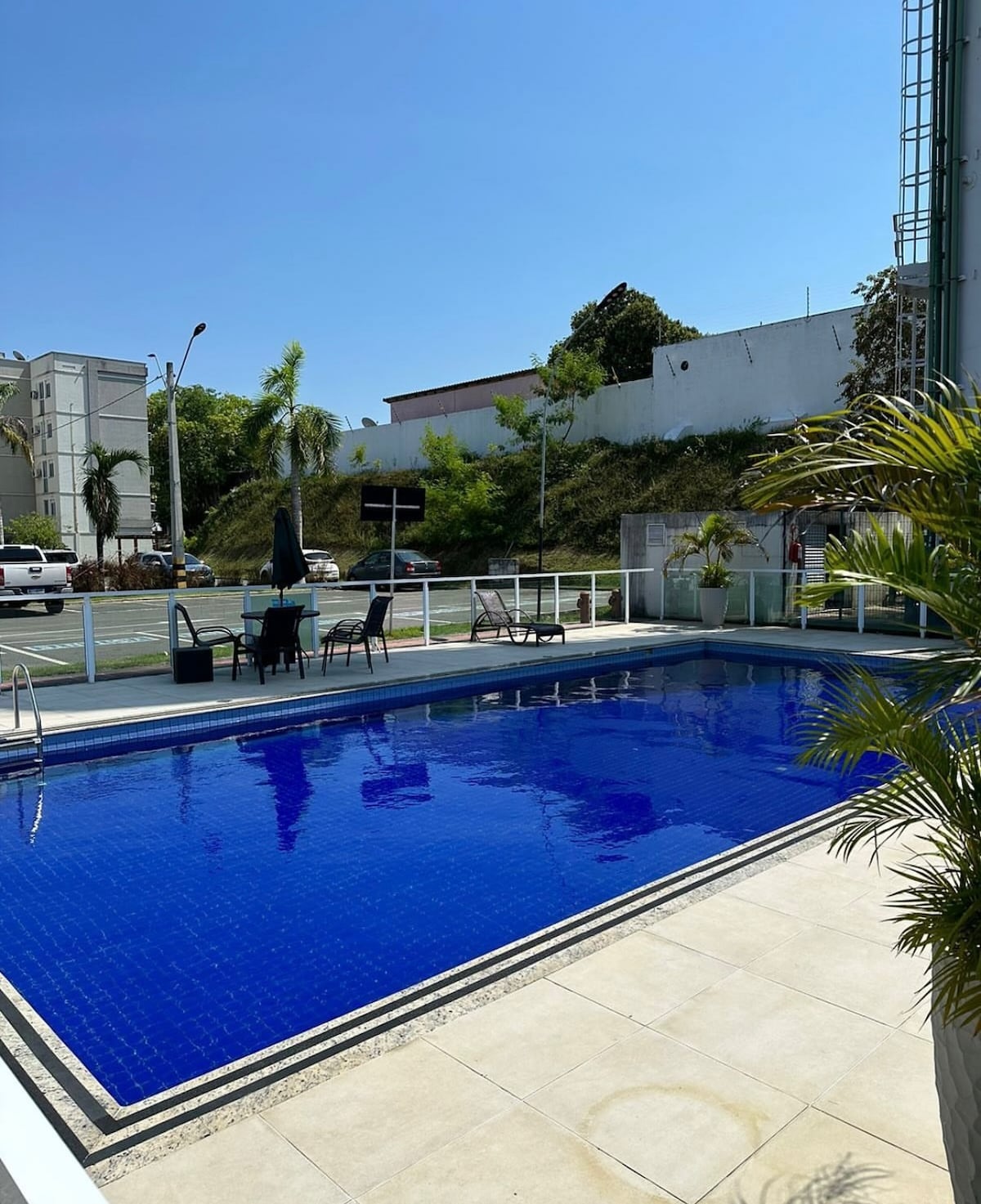 A clear blue pool is surrounded by a textured stone patio. Several patio chairs and tables are positioned nearby, with palm plants adding greenery to the space. Bright sunlight enhances the vibrant colors, and a street can be seen in the background.