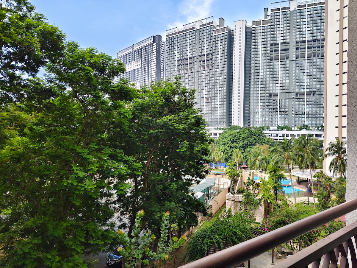 A view from the balcony highlights lush green trees in the foreground, providing a contrast to the towering modern buildings in the background. A glimpse of a pool area can be seen, framed by tropical plants, creating a serene outdoor atmosphere.