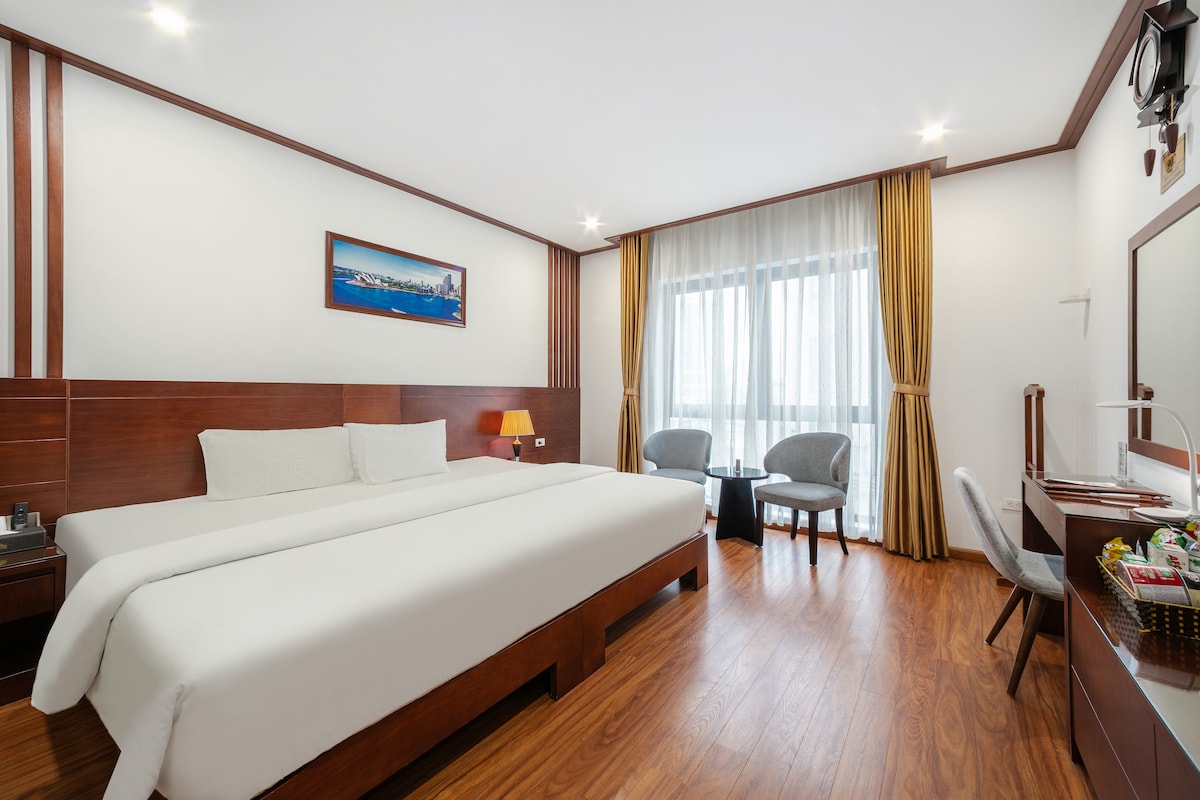 A well-appointed guest room features a spacious bed with crisp white linens, complemented by warm wooden accents. Two armchairs are positioned by the large window adorned with sheer curtains, allowing natural light to fill the room. A small desk with a mirror and decorative items is also present.