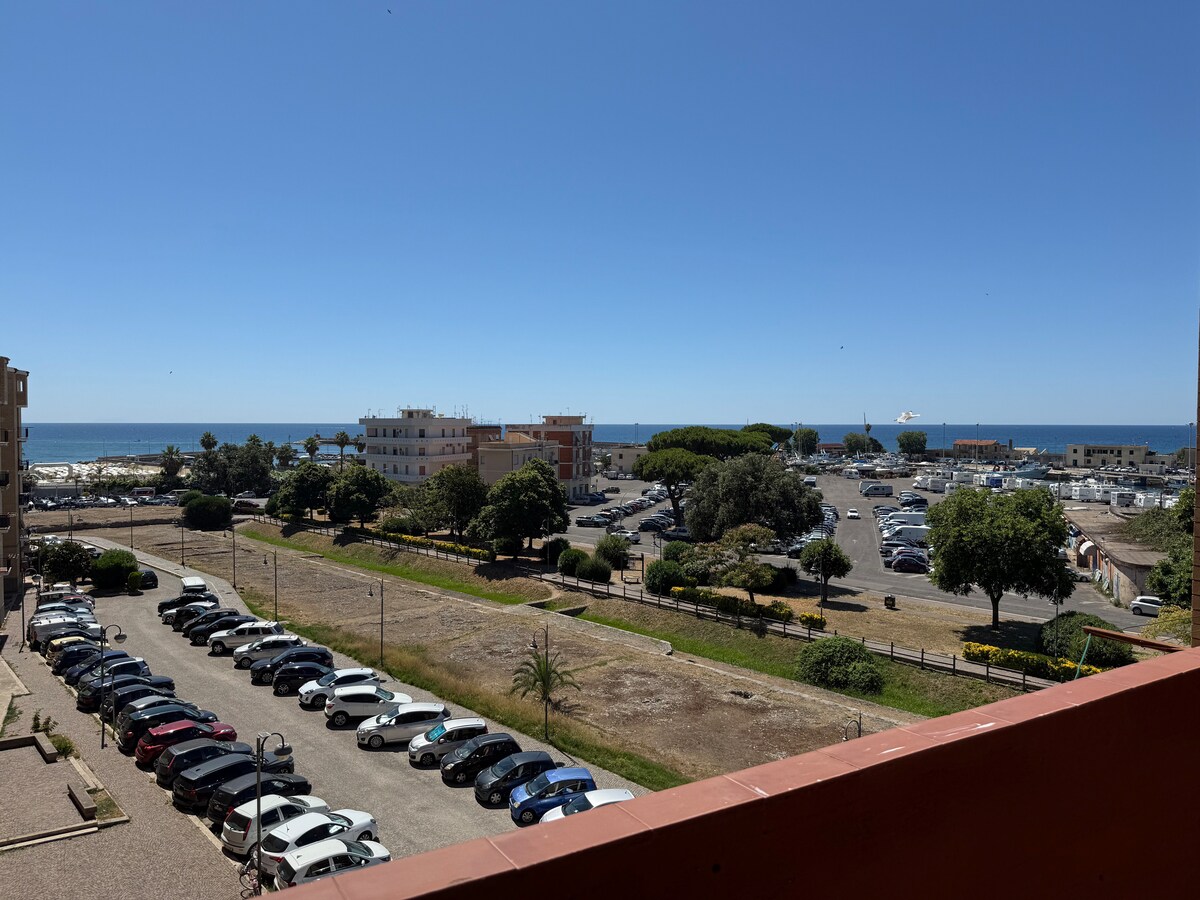 An outdoor view is presented, showcasing a spacious parking area filled with vehicles, alongside lush greenery and coastal scenery. The serene ocean can be seen in the distance, framed by clear blue skies, creating a peaceful backdrop to the residential area.