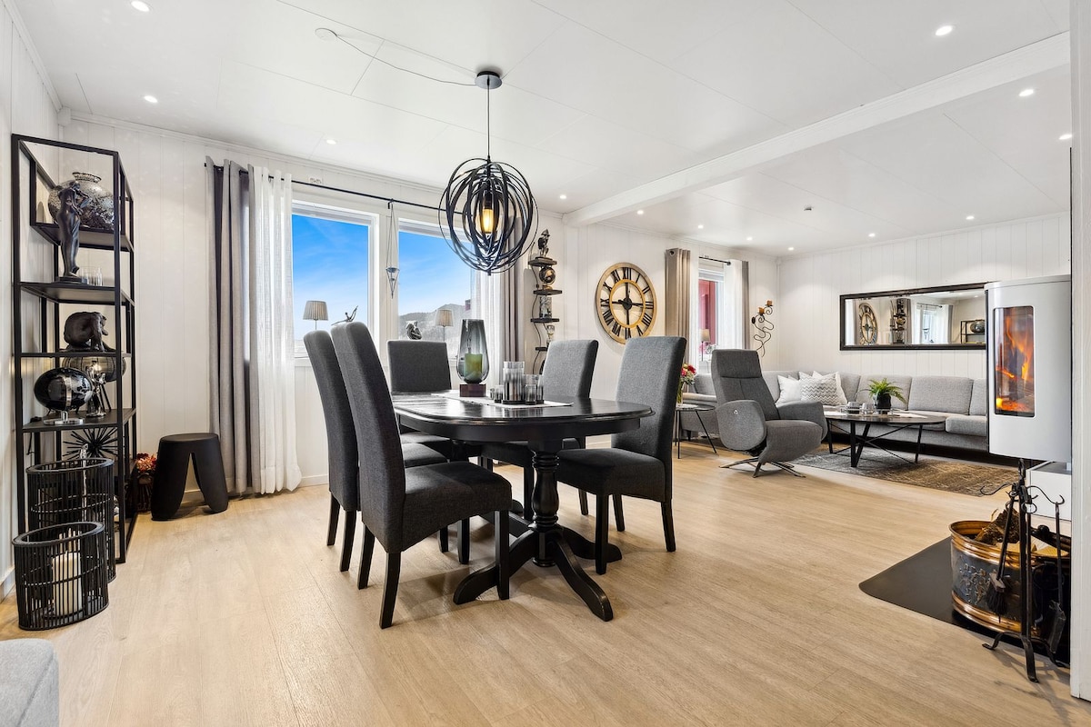 A spacious dining area features a round black table surrounded by six upholstered chairs. A modern light fixture hangs above, and large windows provide natural light. The open living space includes a gray sofa and decorative shelving, creating a welcoming atmosphere.