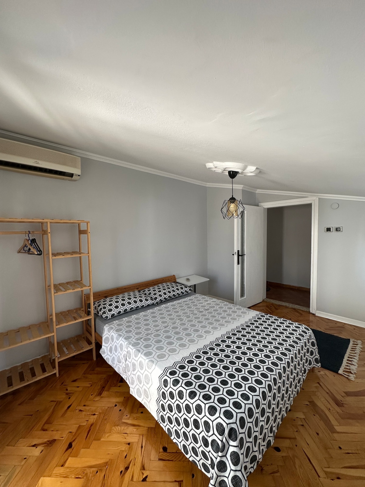 A simple bedroom features a spacious bed dressed in a white sheet with circular patterns. A wooden ladder-style bookshelf stands against one wall, while a small nightstand is positioned beside the bed. Natural light is present, accentuating the wooden floor and neutral wall color.