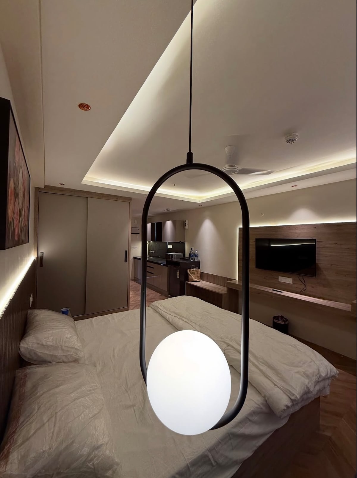 A modern bedroom is illuminated by a distinctive pendant light hanging above the bed. Soft bedding is neatly arranged on the spacious bed, and natural light is subtly filtered through ambient lighting in the room. A sleek TV is mounted on the wall opposite.