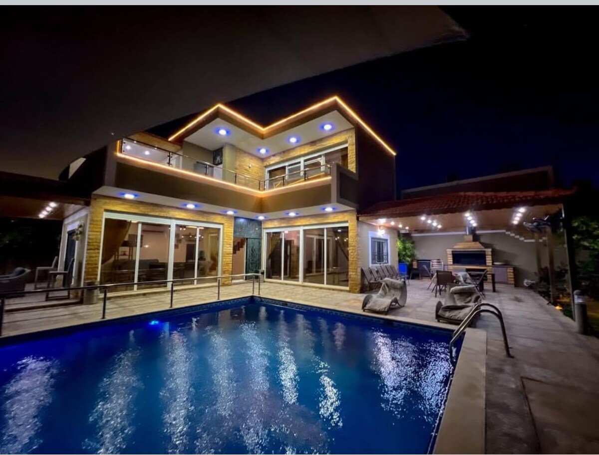 The outdoor area features a well-lit swimming pool, surrounded by lounging chairs. The two-story house is illuminated with decorative lighting, showcasing expansive glass doors that offer views of the interior. A spacious terrace area is present, enhancing the inviting ambiance for relaxation.