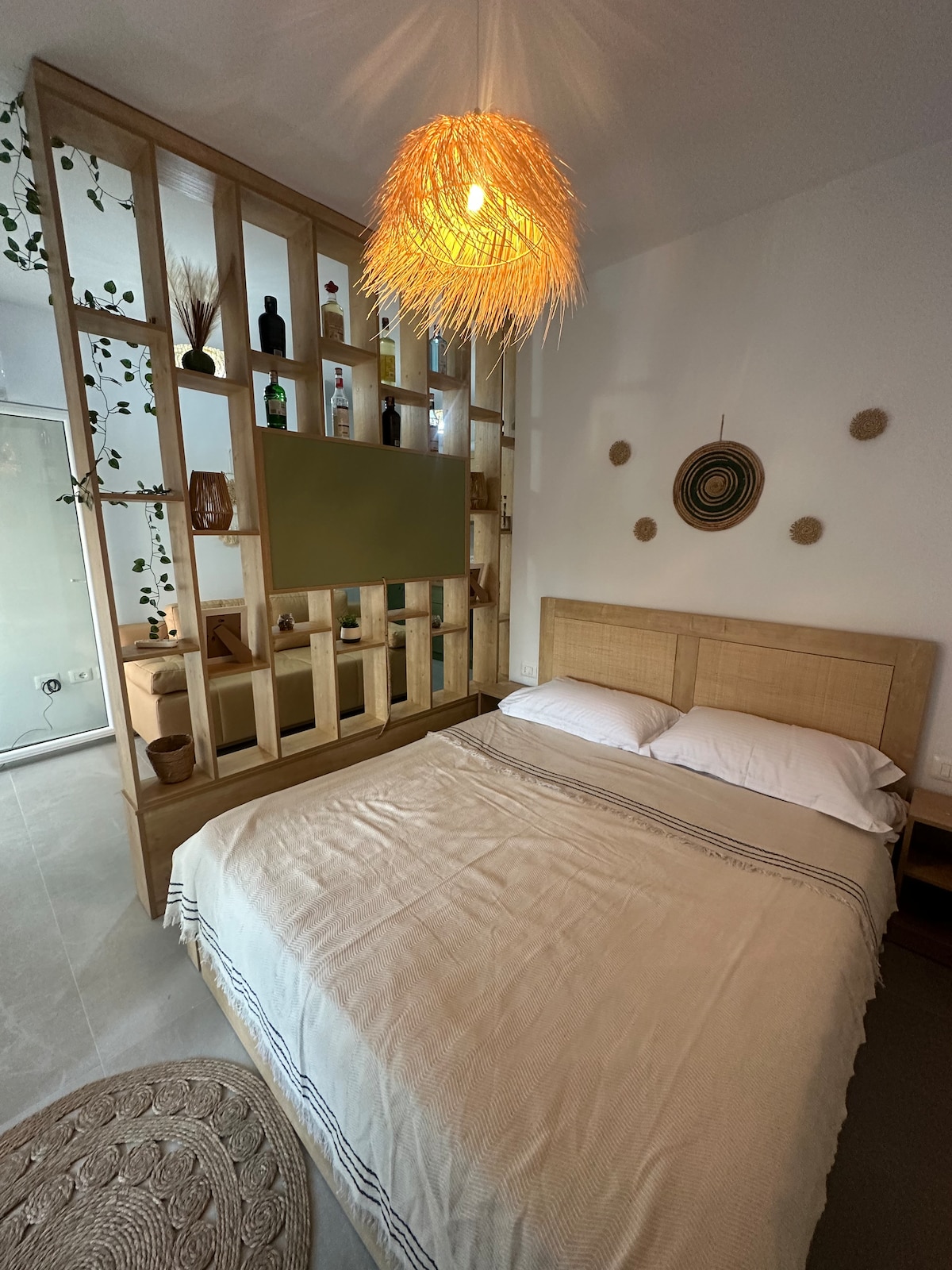 A cozy sleeping space is highlighted by a spacious bed draped in a neutral-colored coverlet. A decorative wooden partition with plant accents and glass shelves displays various items. Soft lighting is provided by a woven pendant lamp above, enhancing the serene ambiance.