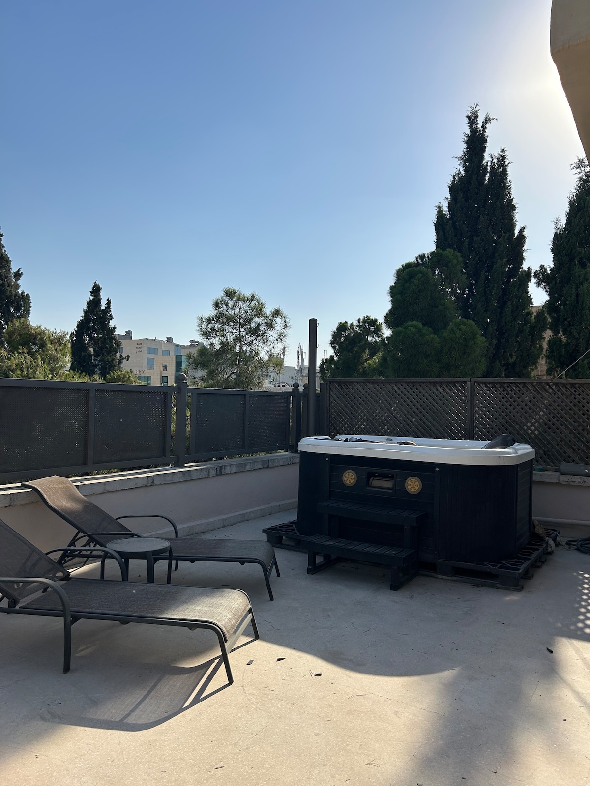 A private rooftop space features a jacuzzi surrounded by a wooden lattice fence, complemented by two lounge chairs. Sunlight illuminates the area, and greenery from surrounding trees adds a natural touch to the urban setting.