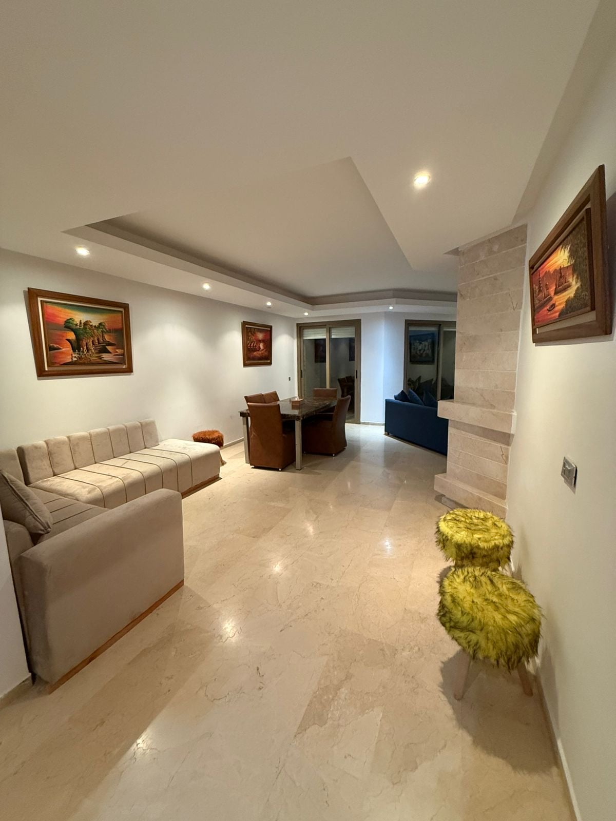 An open-concept living area features a light-colored sofa, paired with two textured stools. A dining table with four chairs is visible, accompanied by artwork on the walls. Soft lighting highlights the marble flooring, while a doorway leads to another room.
