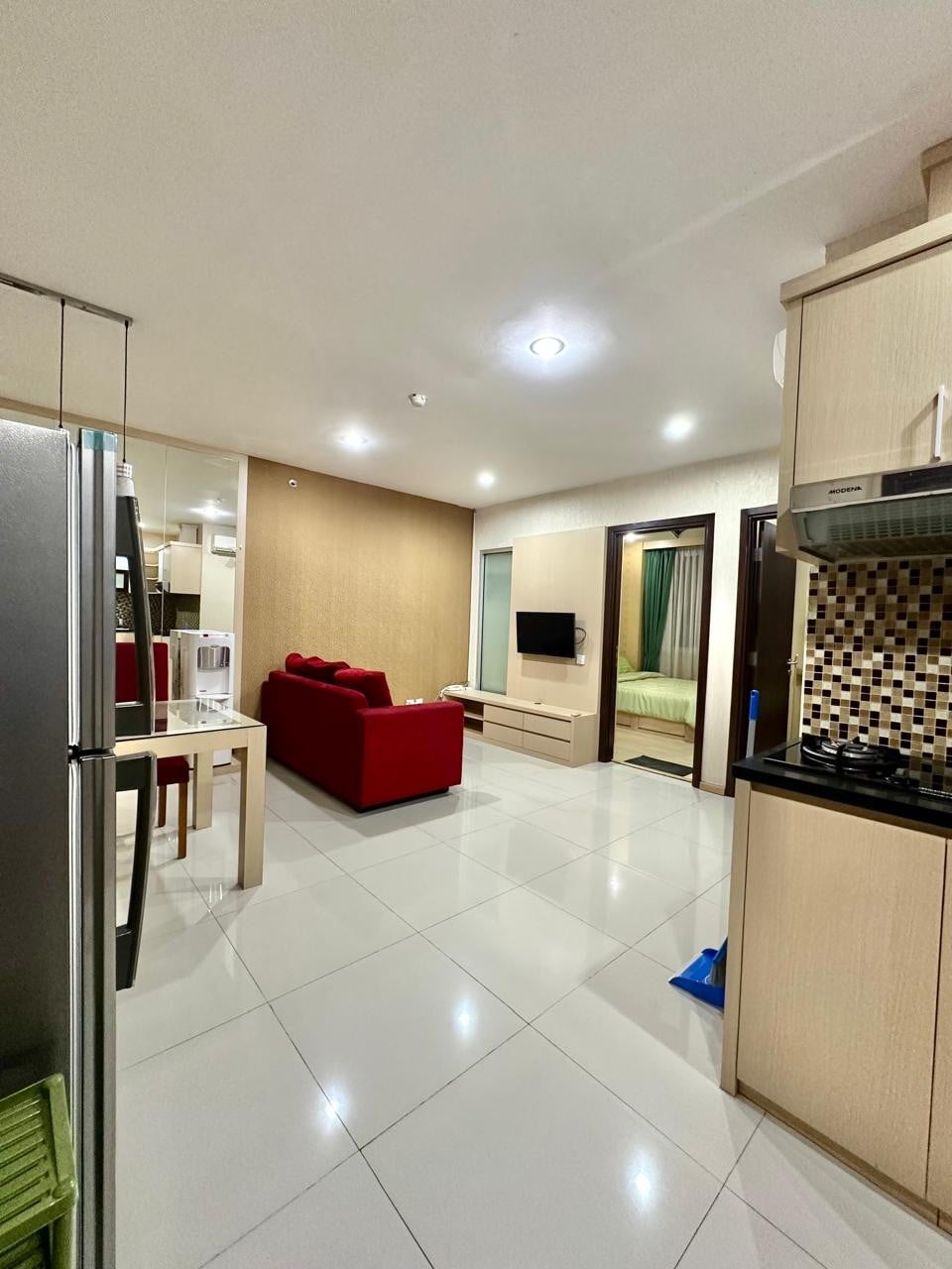 A modern living space is visible, featuring a red sofa positioned against a light wooden wall. A compact dining table is set nearby, and a flat-screen TV is mounted on the opposite wall. A glimpse of a sleeping area is seen through partially open doors, with kitchen elements off to the side.