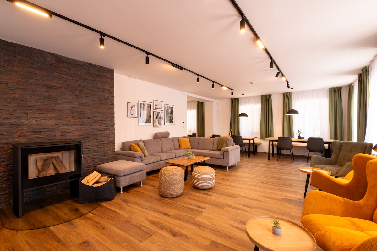 A spacious common area is highlighted by a stone fireplace and a variety of seating options. Soft lighting creates a cozy environment. Large windows allow natural light to illuminate the room, which features wooden flooring and a selection of decorative elements.