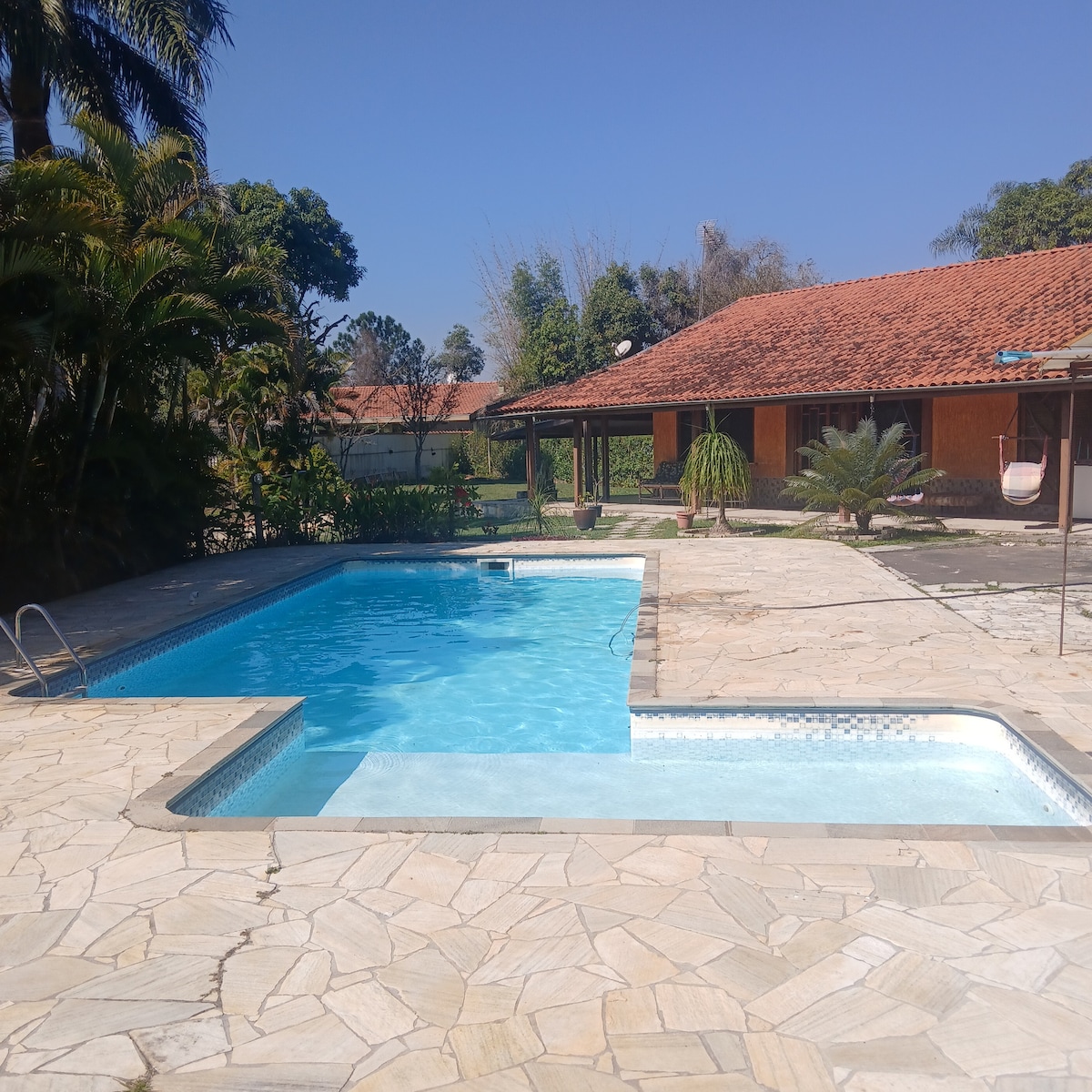 A spacious outdoor area is shown, featuring a large swimming pool with a shallow section and surrounded by paved stone. Lush greenery is visible, alongside a charming house with a red-tiled roof in the background, offering a welcoming environment for relaxation.