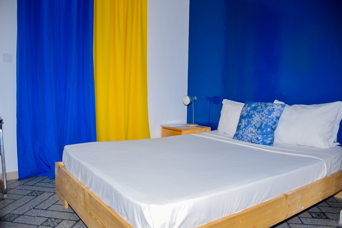 A bright sleeping area features a spacious bed with white linens, accented by a decorative blue pillow. Blue and yellow curtains frame a nearby window, and a small bedside table holds a lamp, contributing to the overall clean and simple design.