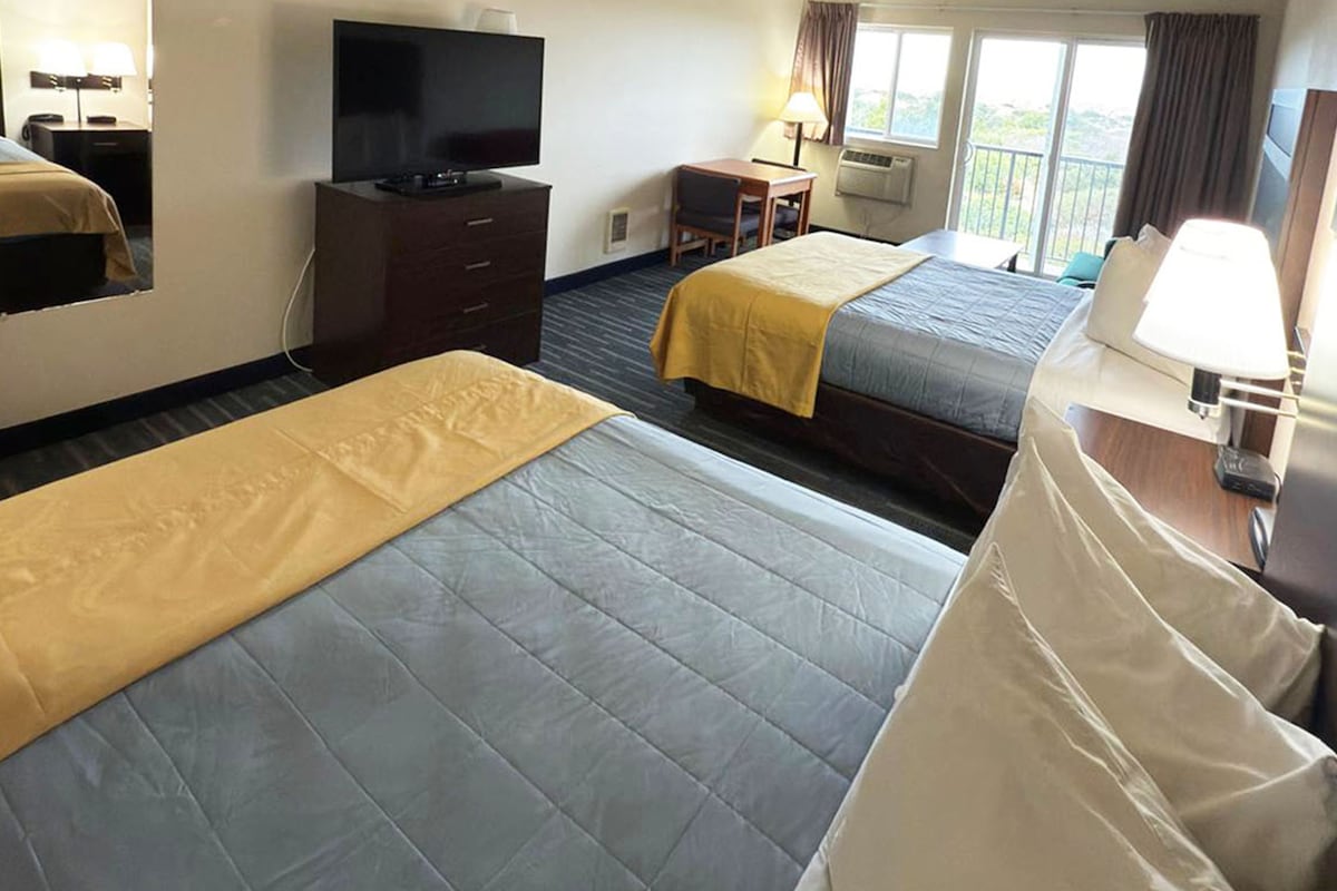 A spacious hotel room is presented with two beds, one covered in a yellow blanket and the other in a light grey coverlet. A flat-screen TV is mounted on the wall opposite, and a wooden table with a chair is visible near the window, which offers natural light.