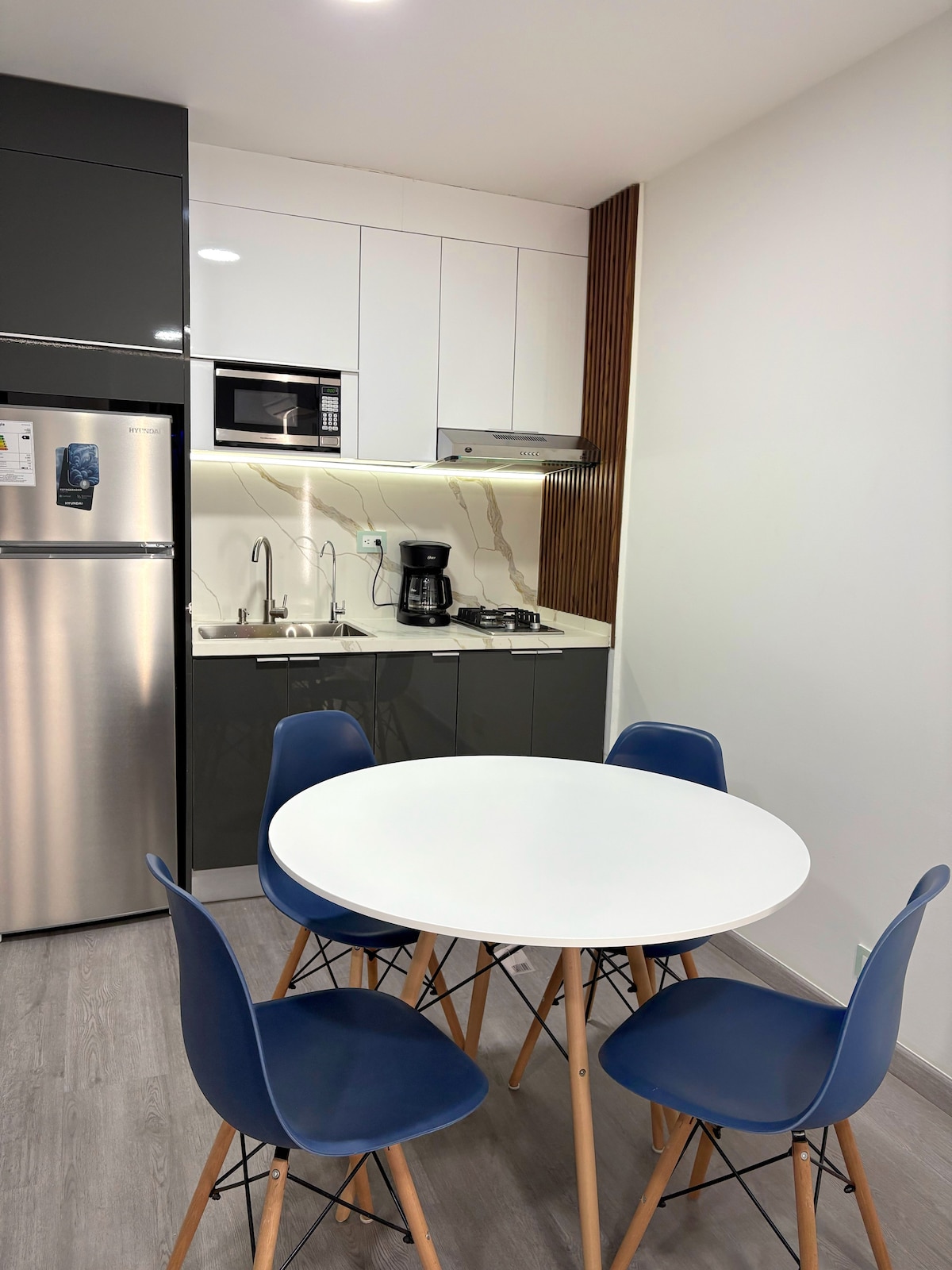 A modern kitchenette is presented with sleek cabinetry and essential appliances, including a microwave and a coffee maker. A round dining table is surrounded by four chairs, creating a functional space for enjoying meals. Ample light illuminates the area, highlighting its clean lines.