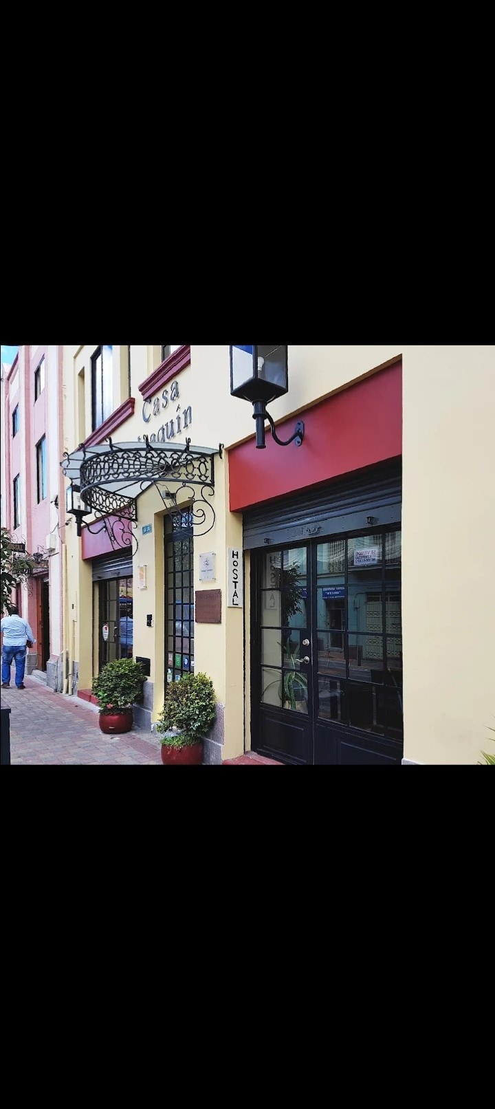 The exterior of the hotel features a charming façade with a welcoming sign above the entrance. Large glass doors showcase the inviting interior. Potted plants are arranged alongside the entrance, enhancing the aesthetic appeal of the building, framed by a vibrant yellow wall.