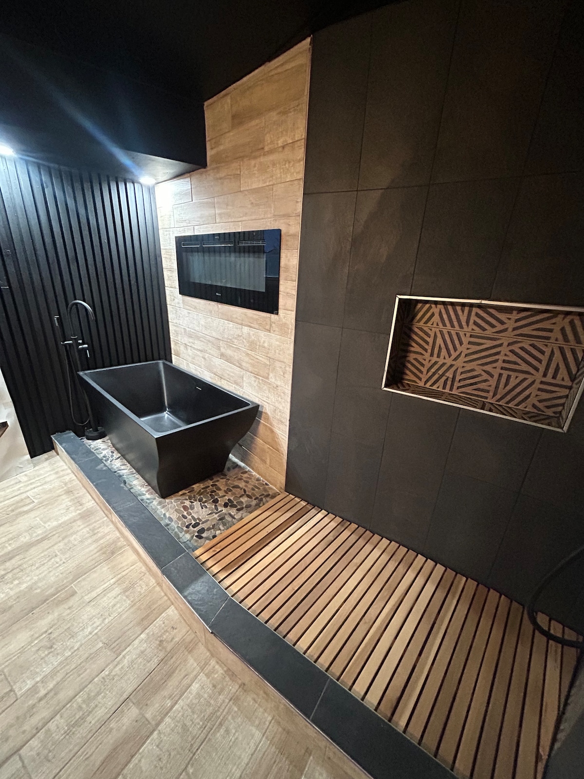 A modern bathroom space is showcased, featuring a black freestanding bathtub positioned against a textured wall. The floor consists of wooden slats adjacent to a patterned tile inset. Natural wood accents and soft lighting contribute to an inviting ambiance.