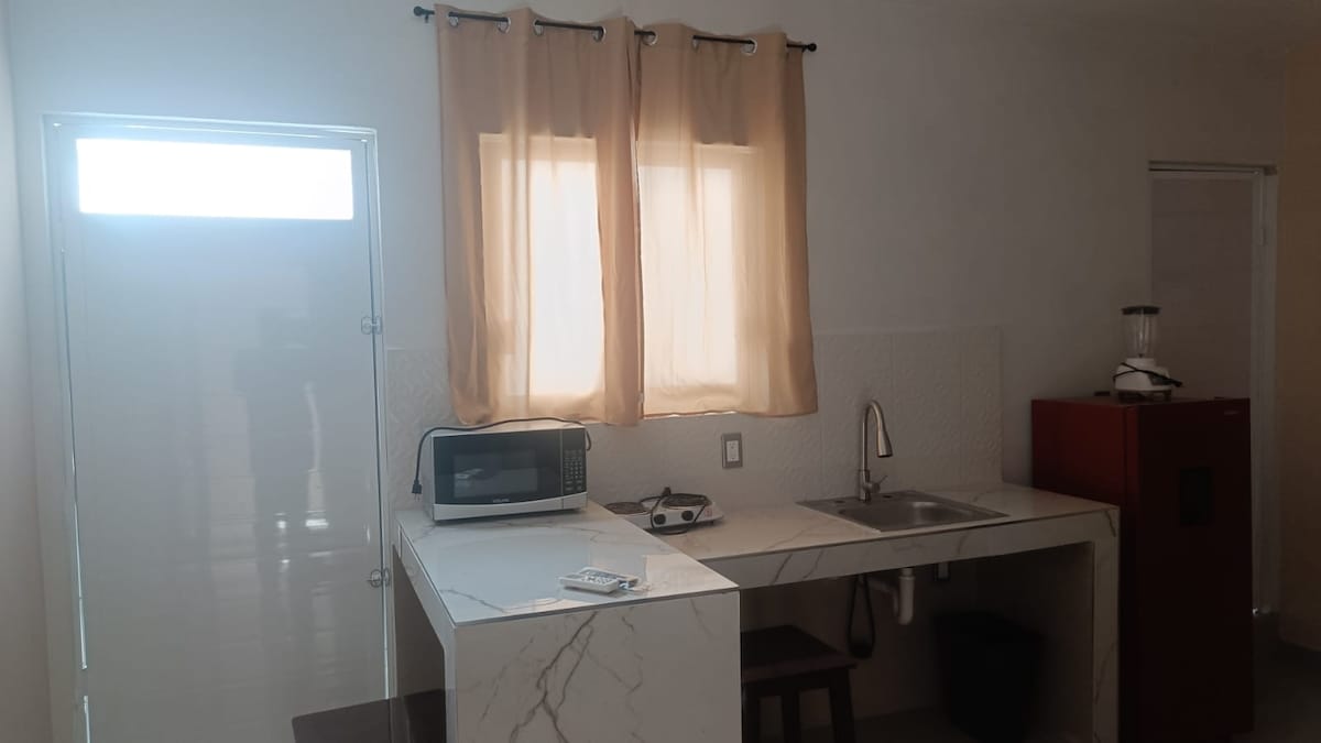 The kitchen area features a marble-topped counter with a microwave and a small cooking surface. Light enters through a window covered by sheer curtains, illuminating the space. A dark cabinet is positioned to the right, providing additional storage.