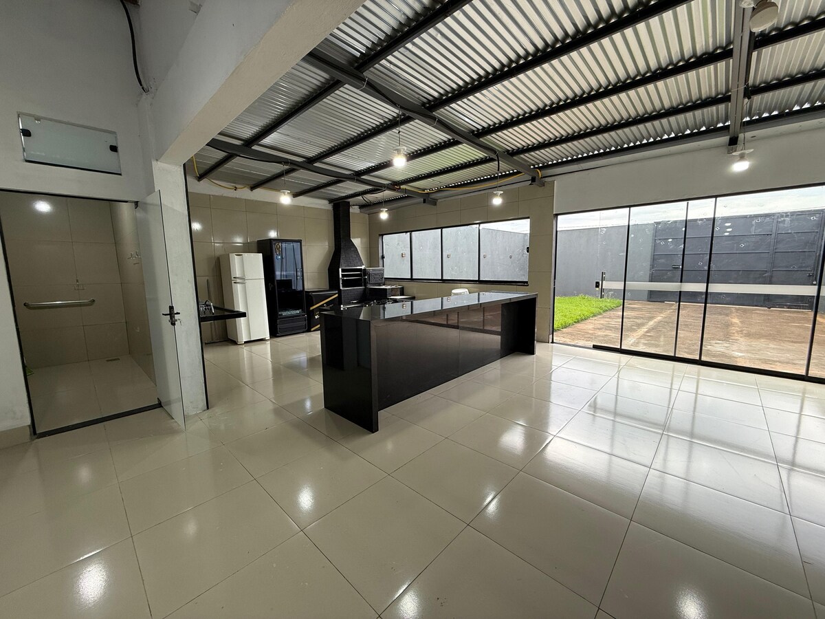 A spacious kitchen area features a modern design with glossy cabinetry and a central island, complemented by a large refrigerator and a built-in grill. Expansive glass doors allow natural light and a view of the outdoor space, enhancing the openness of the area.