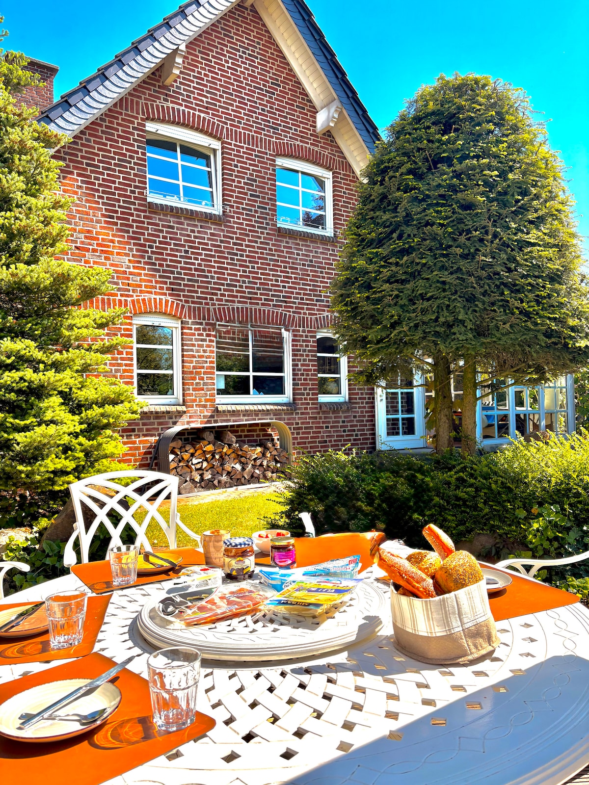 A charming outdoor dining area is set with a round table, surrounded by a white, intricately designed metal framework. The table features a spread of food items and drinks, complemented by lush greenery and a rustic brick exterior in the background.