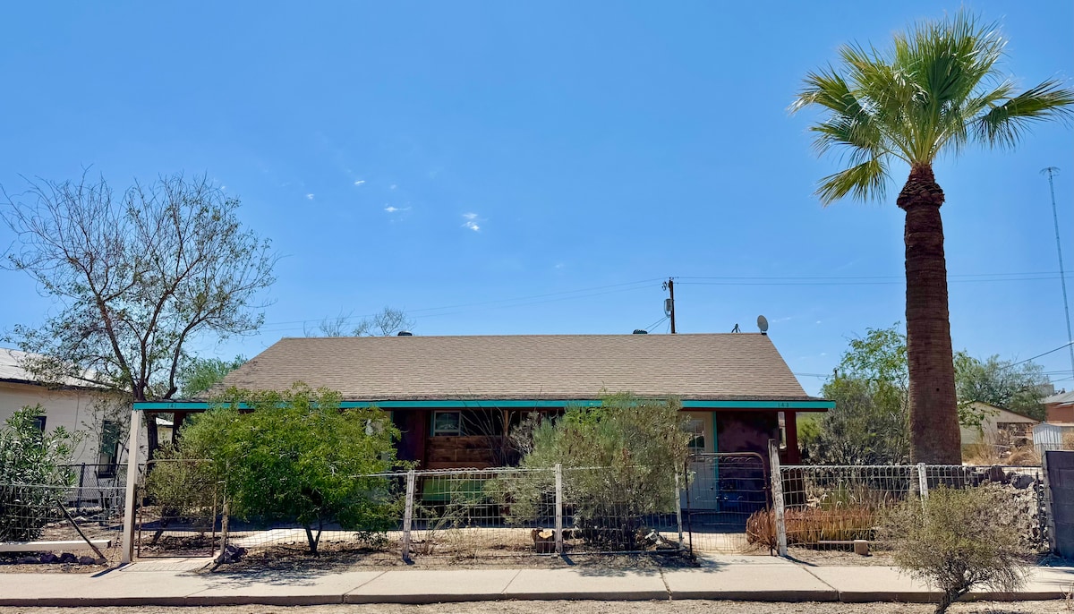 A single-story duplex is shown surrounded by desert landscaping. A tall palm tree stands to the right, complementing the home’s traditional architecture. The exterior features earthy tones and a roof adorned with a simple overhang, while a low fence and native shrubs enhance the outdoor space.
