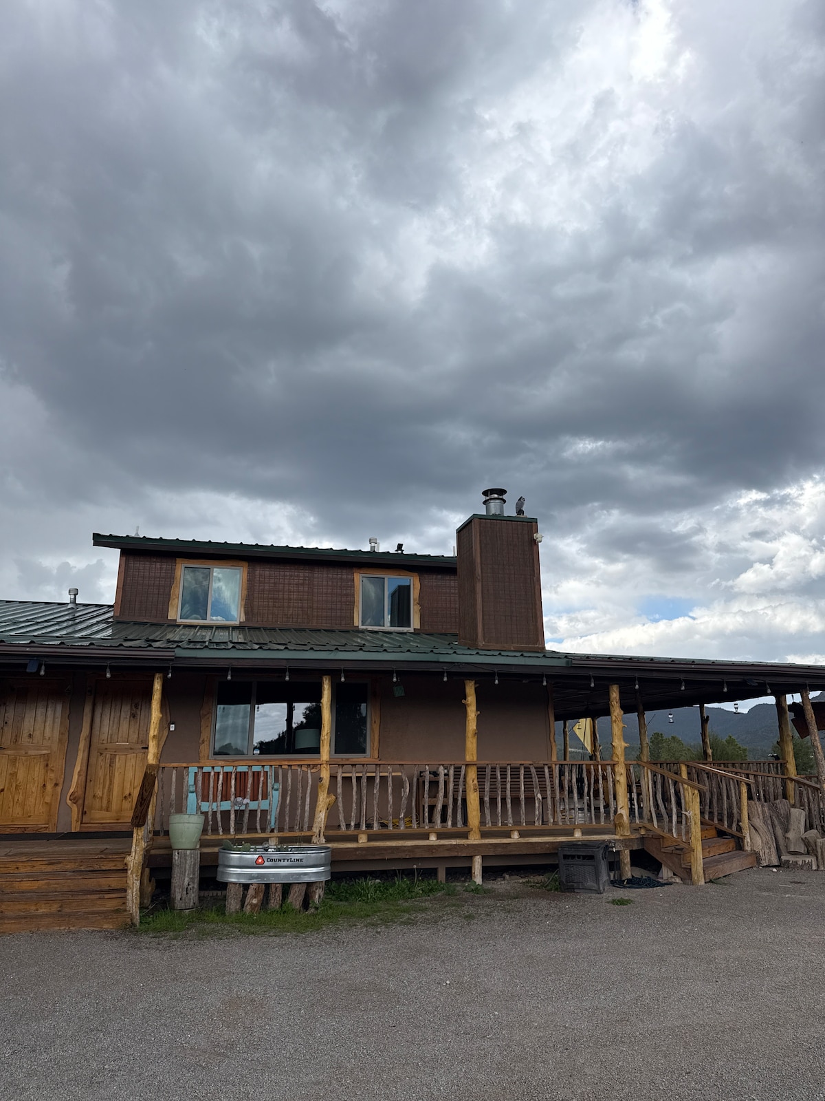 The exterior of the condo is shown, featuring a rustic design with wooden beams and a spacious deck. Large windows provide natural light, while a chimney is visible. A cloudy sky adds a dramatic backdrop to the mountain retreat.