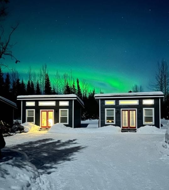 Two cozy cabins are illuminated under a vibrant northern lights sky, contrasting with the deep blue backdrop. Snow blankets the ground, and tall trees stand quietly in the background, creating a serene winter landscape.