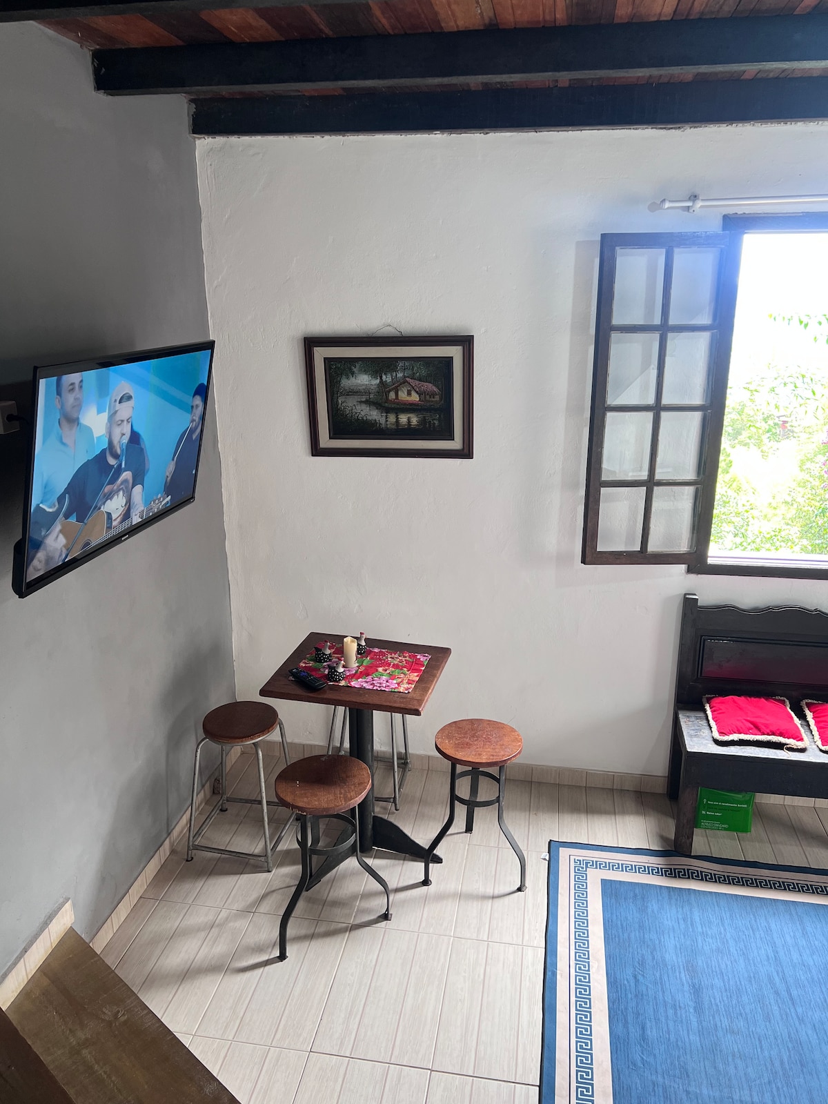 The living area features a small table accompanied by three stools, positioned beneath a wall-mounted TV. A framed artwork adorns the wall, while a window allows natural light to illuminate the space, which is complemented by a blue area rug on the floor.