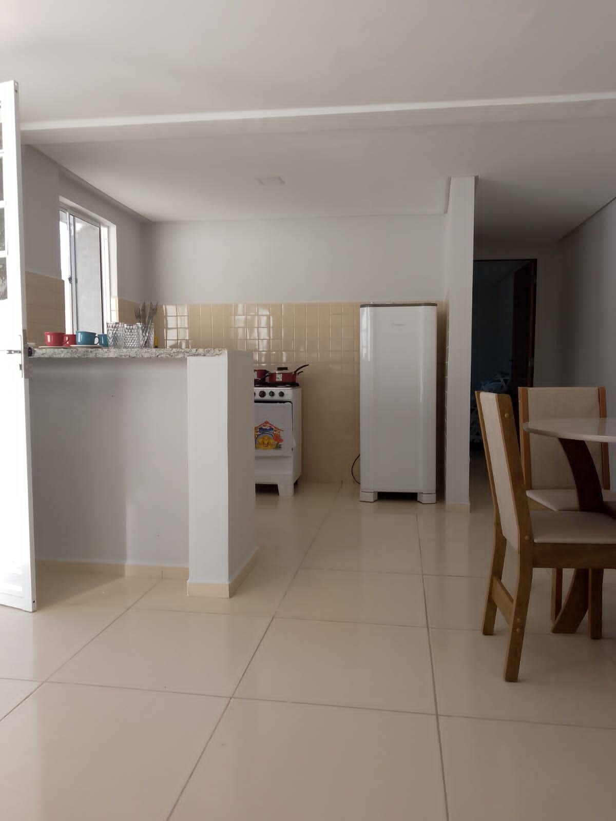 A clean and functional kitchen area features a white refrigerator and a small stove with a red and white design. Natural light floods the space through a window, highlighting beige tiles on the walls and floor. A dining table with wooden chairs is situated nearby.