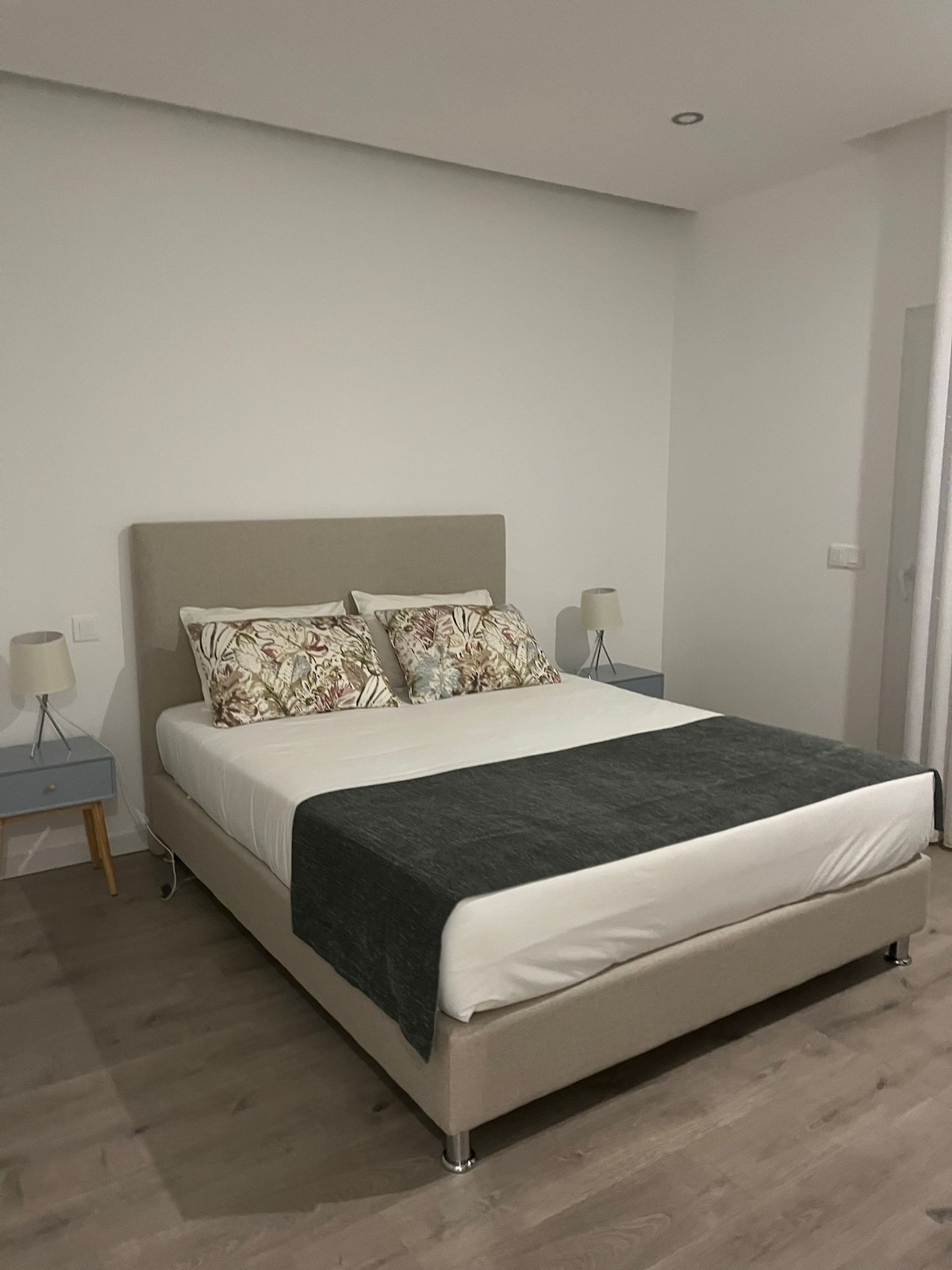 A spacious bedroom features a comfortable bed with decorative pillows and a soft throw. Two bedside tables with matching lamps are placed on either side of the bed. The light wooden flooring complements the neutral wall colors.