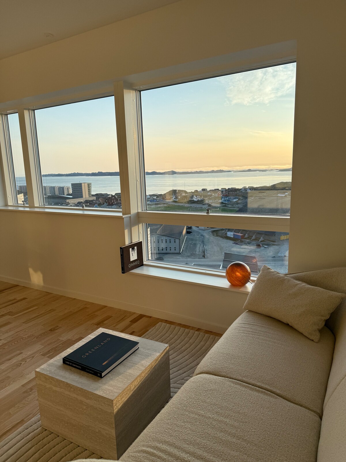 A cozy seating area features a light-colored sofa positioned near large windows, offering sweeping views of the fjord and city. A small table holds a book, and a decorative object sits atop the table, illuminated by natural light streaming in from the windows.