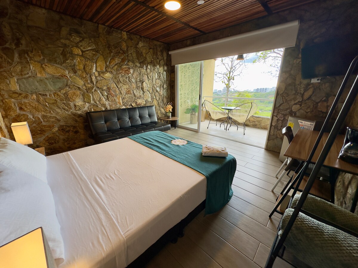 A cozy room features a spacious bed with a soft white blanket, complemented by rustic stone walls and wooden accents. A sitting area with a sleek black sofa overlooks an outdoor balcony with scenic views. Natural light fills the space, enhancing the comfortable ambiance.