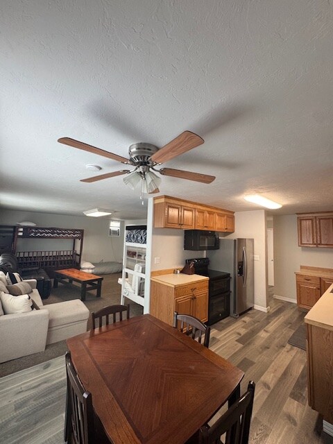 An open living area features a kitchen with wooden cabinets and stainless steel appliances. A dining table with four chairs is positioned near the kitchen. A comfortable sofa and futon bunk beds are visible in the background, with a ceiling fan providing airflow.