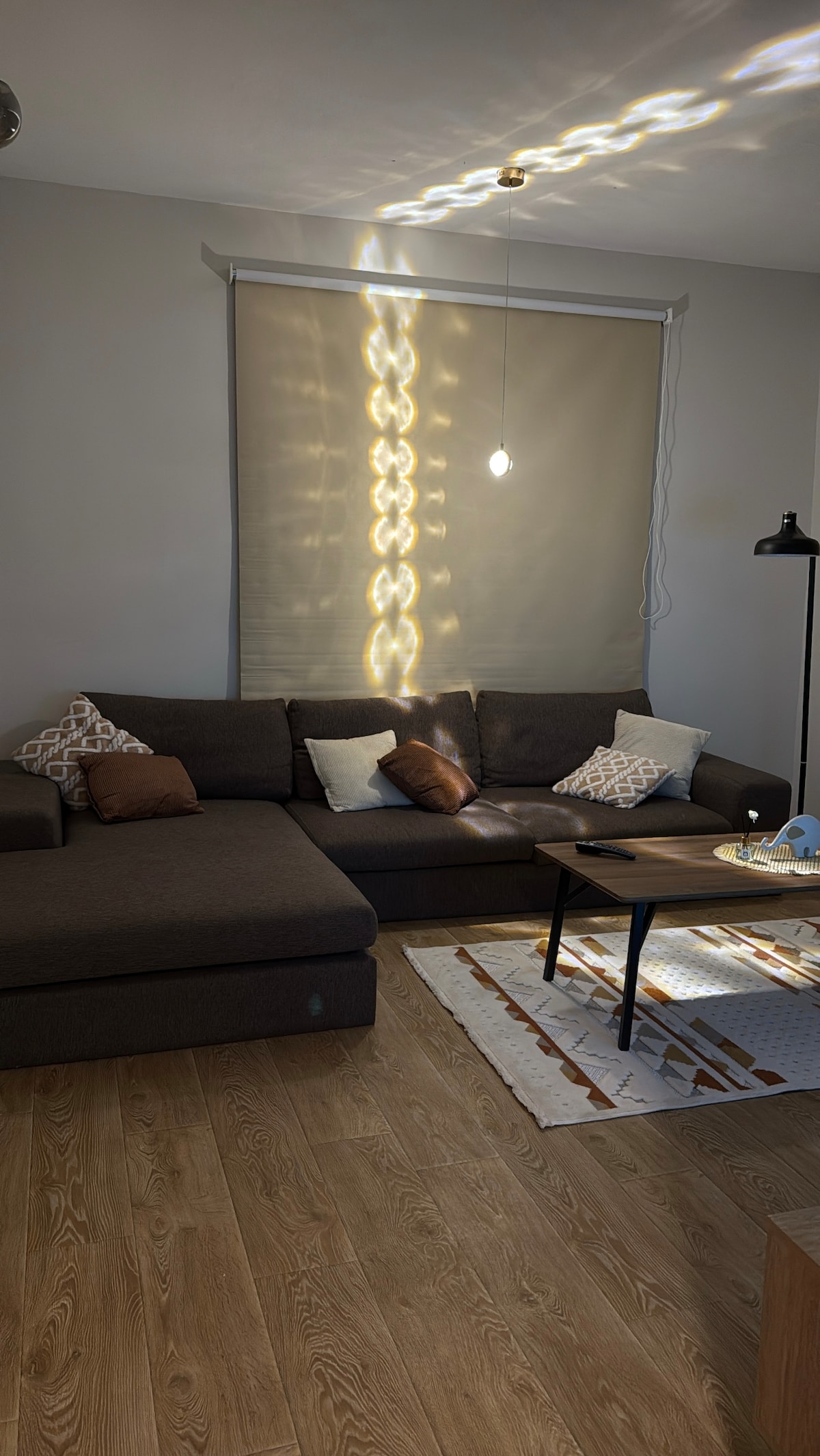 A cozy seating area is featured with a dark sectional couch adorned with decorative pillows. A small coffee table sits in front, accompanied by a decorative rug. Natural light streams in, creating patterns on the wall from a nearby window treated with a light-filtering shade.