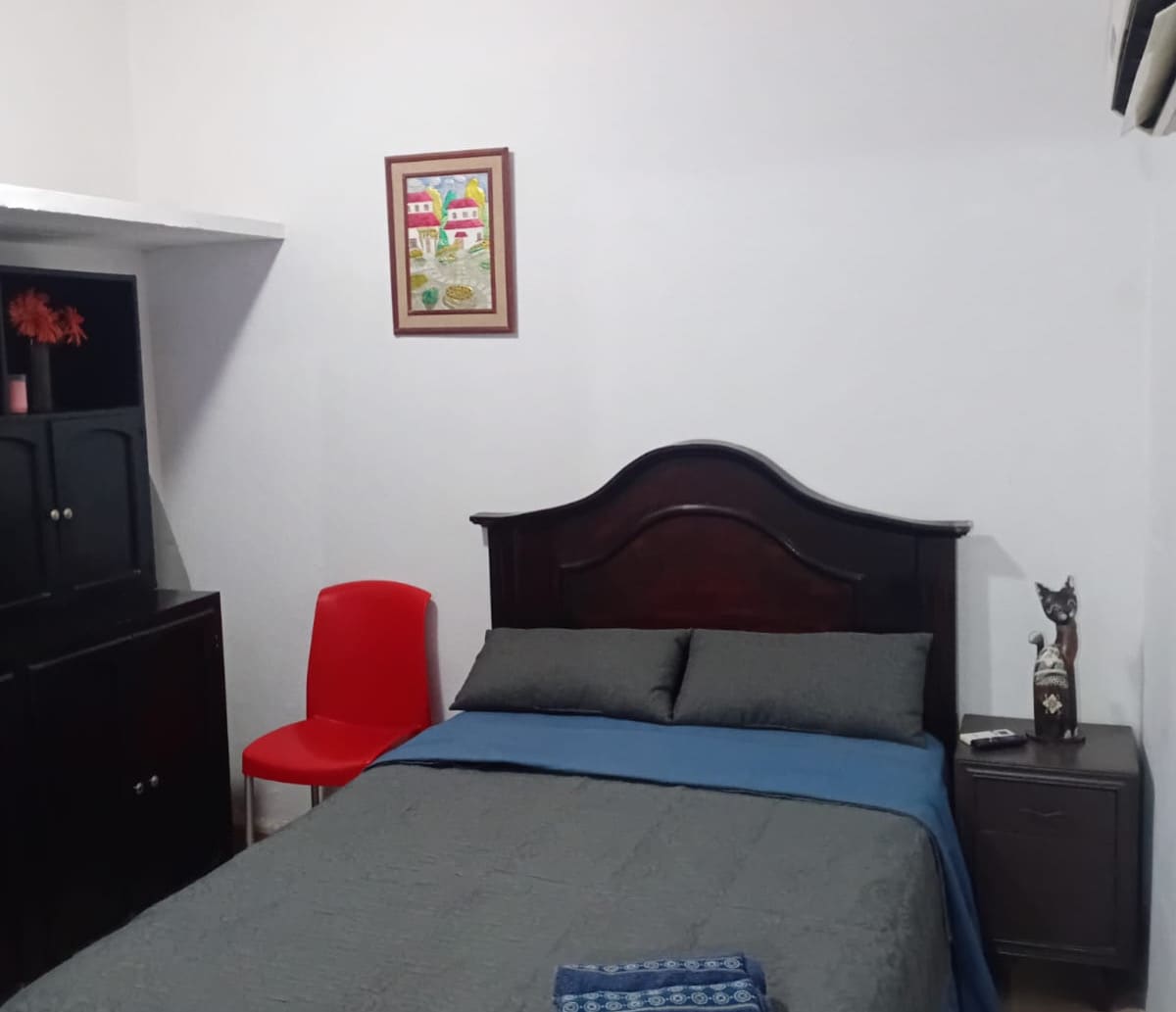 The sleeping area features a spacious bed with a dark wooden headboard, adorned with grey bedding. A vibrant blue throw lies on the bed. A red chair and a decorative piece can be seen in the corner, beside a bedside table with a small figurine.