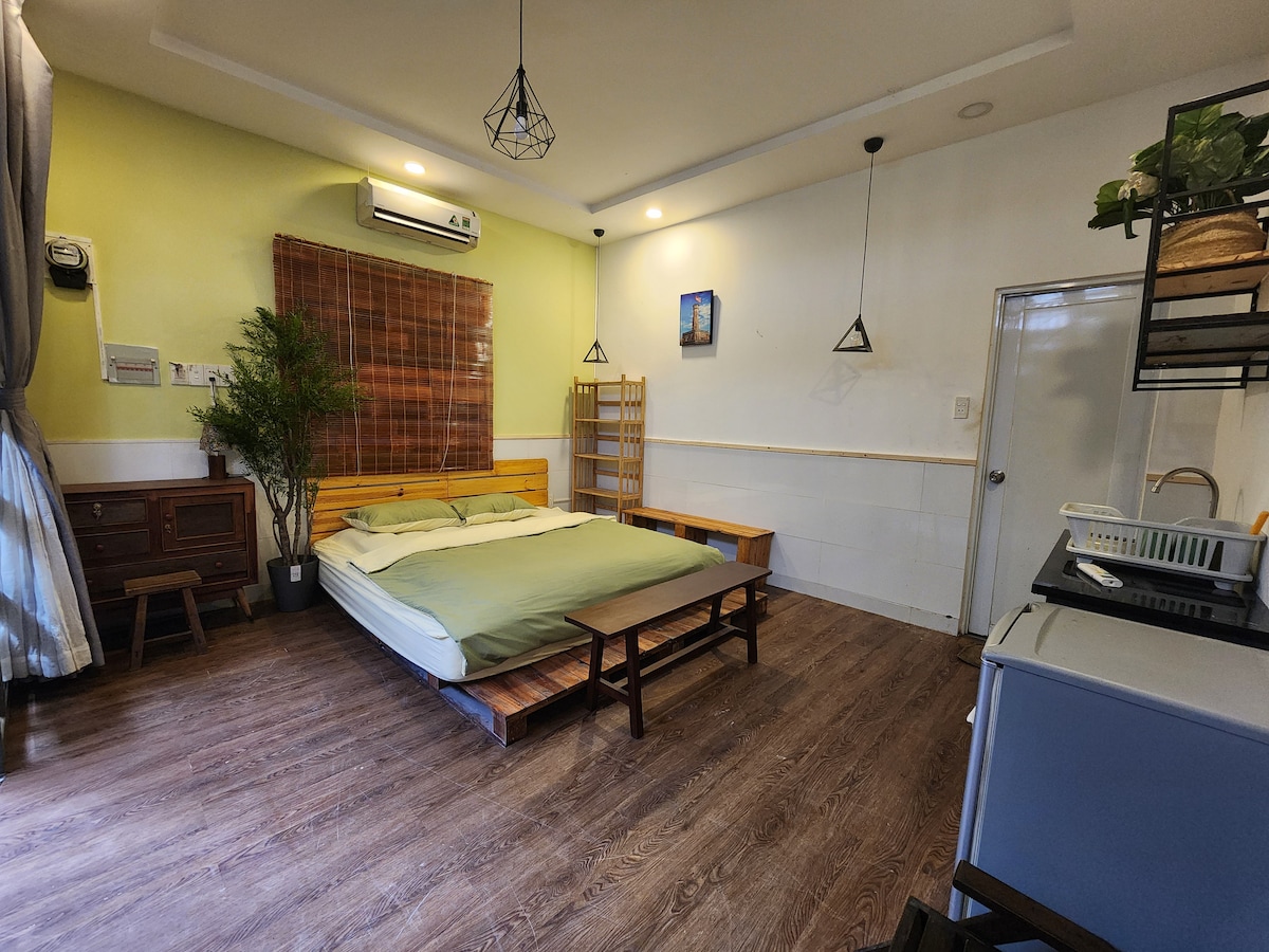 A minimalist bedroom is featured with a spacious bed topped with a soft green blanket. Wooden furniture, including a low table and a shelf, enhances the simplicity. Natural light enters through a window, while air conditioning is provided for comfort.