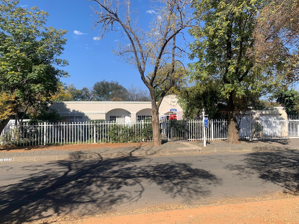 The exterior of the property is framed by a white picket fence, surrounded by leafy trees casting soft shadows. The building features arches above the windows, with pathways leading to the entrance, all set amidst a serene, residential neighborhood.