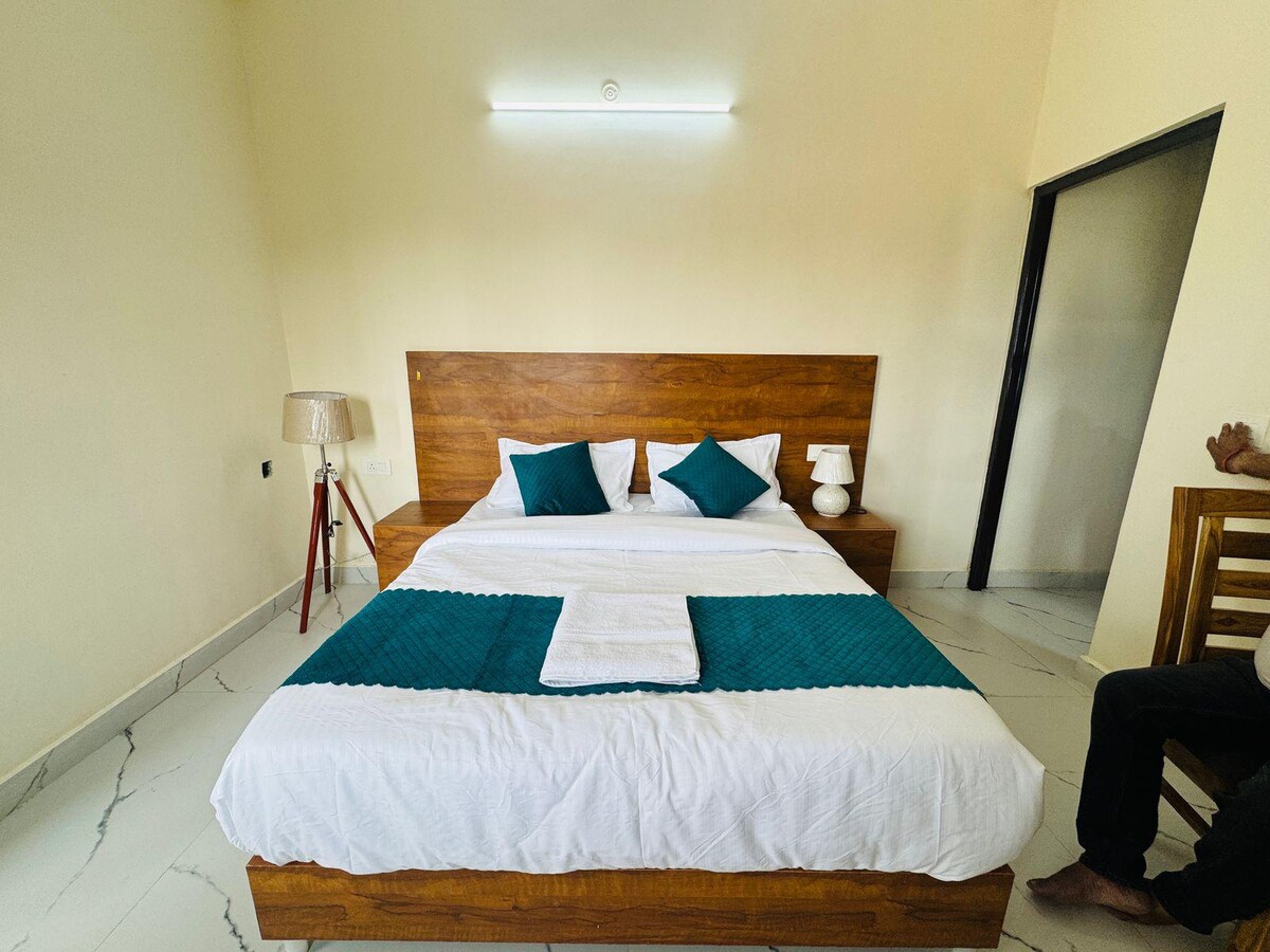A bright sleeping area features a wooden bed with a teal accent blanket and decorative pillows. A small lamp stands beside the bed on a wooden nightstand, while a chair is positioned next to the wall. Natural light fills the space through the open door.