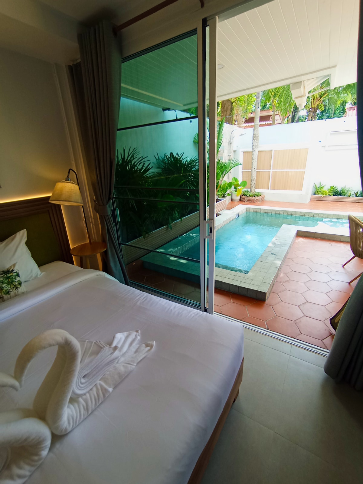 A serene bedroom scene features a spacious bed with soft linens and a decorative pillow. Glass doors open to a private saltwater plunge pool surrounded by greenery. Natural light enhances the inviting atmosphere, with palm trees visible through the glass.