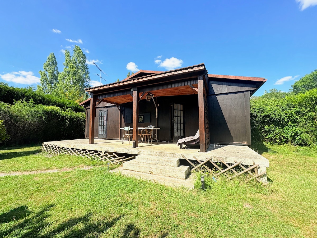 The chalet is set on a green lawn, featuring a wooden deck with steps leading up to it. Outdoor furniture is arranged under a covered area, and large windows enhance the cozy appearance. Surrounding vegetation adds a natural backdrop to the peaceful setting.
