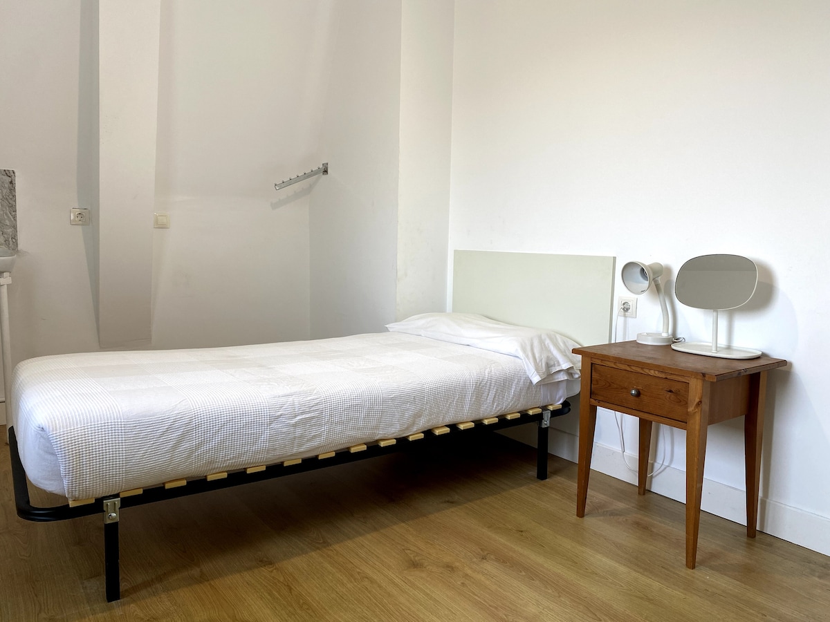 A single bed with a simple white bedding is positioned against a bright wall. A wooden bedside table supports a mirror and a small lamp. Natural light fills the room, highlighting the light wooden flooring.