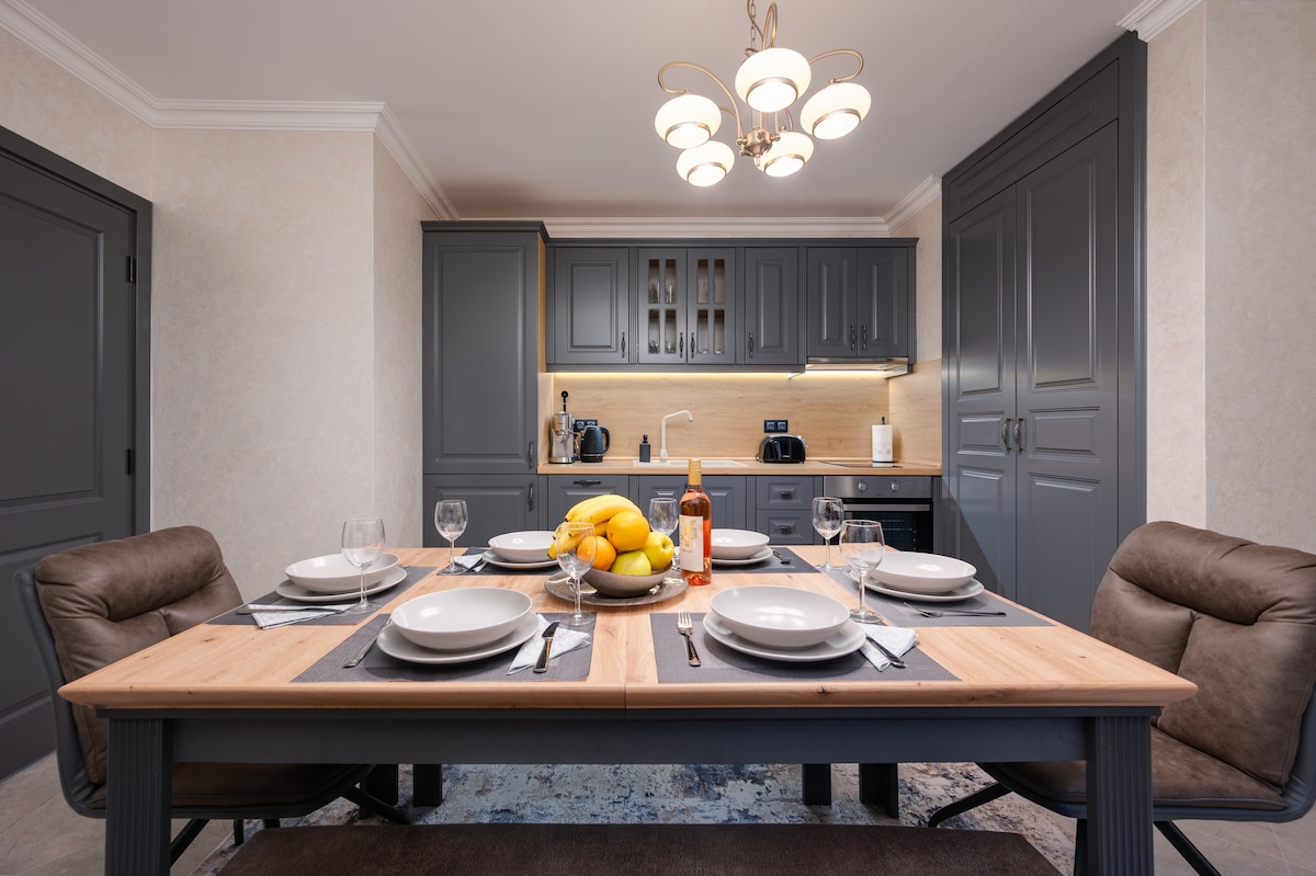 A modern kitchen is presented with dark cabinetry and contemporary lighting. A dining table is set with plates, glasses, and a centerpiece of fresh fruit. Kitchen appliances can be seen in the background, complemented by warm wooden accents.