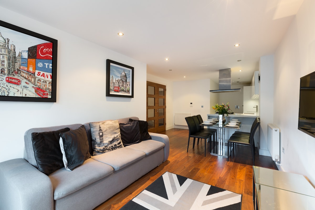An open-plan living area features a comfortable gray sofa adorned with a decorative throw. A dining table with black chairs is positioned nearby, complemented by modern artwork on the wall. Natural light illuminates the space, showcasing the warm wooden floor.