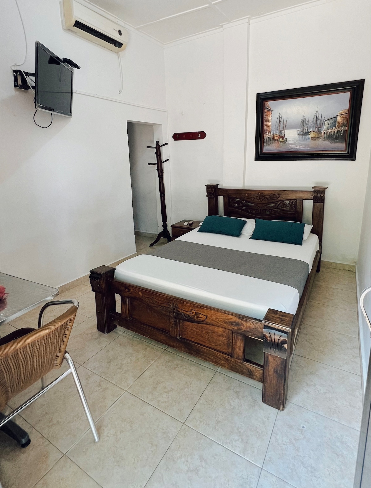 A simple room features a wooden bed with a gray and white spread. A small dining table with two chairs is visible, along with a wall-mounted television. A painting depicting a cityscape hangs above the bed, and a coat rack stands in the corner.