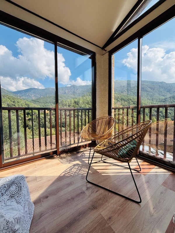A sunlit corner features two woven chairs with a mountainous view visible through expansive glass windows. The natural landscape includes lush greenery and soft clouds, creating a serene atmosphere. Wooden flooring complements the space, enhancing the connection to nature.