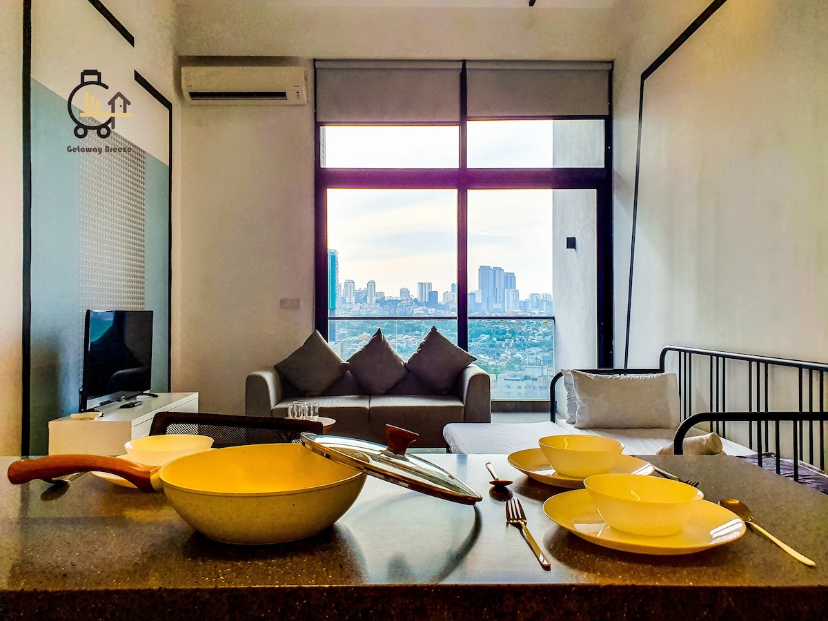 A modern kitchen area is showcased with tableware arranged neatly. A spacious living room, including a couch, is visible in the background, with large windows that offer a view of the city skyline. Natural light filters softly into the space.