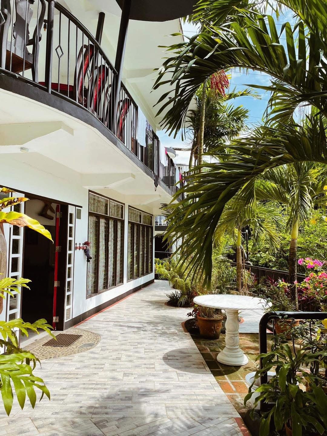 A path lined with tropical plants leads to a white table surrounded by greenery. The two-story building showcases a balcony with black railings and large windows that view the vibrant garden. Sunlight filters through the leaves, creating a peaceful outdoor setting.