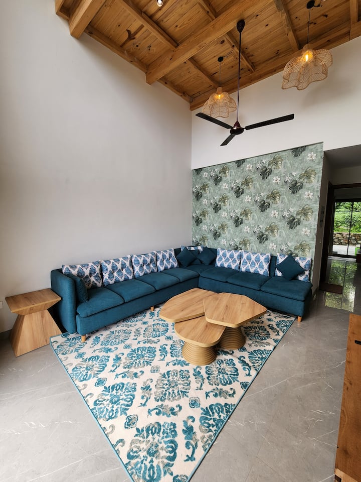 พื้นที่นั่งเล่นกว้างขวางของวิลล่าออกแบบมาเพื่อการพักผ่อน พร้อมเพดานไม้สูง แสงไฟนุ่มนวล และโซฟาสีเขียวอมฟ้าที่อบอุ่นเหมาะสำหรับการผ่อนคลายกับหนังสือหรือการแบ่งปันเสียงหัวเราะกับคนที่คุณรักผ่านเกมกระดาน 